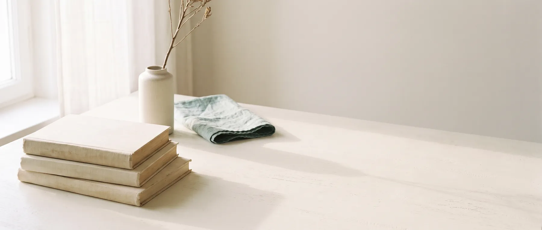 Stacked books with blank covers, a pale ceramic vessel, and teal linen