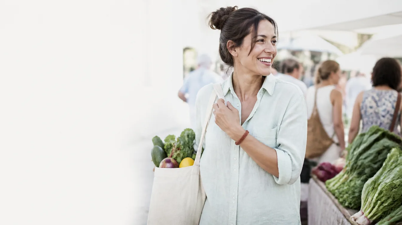 A woman in her early 40s with natural realistic skin laughing while carrying a canvas tote of fresh produce at an outdoor farmers market, standing beside a stall with leafy greens