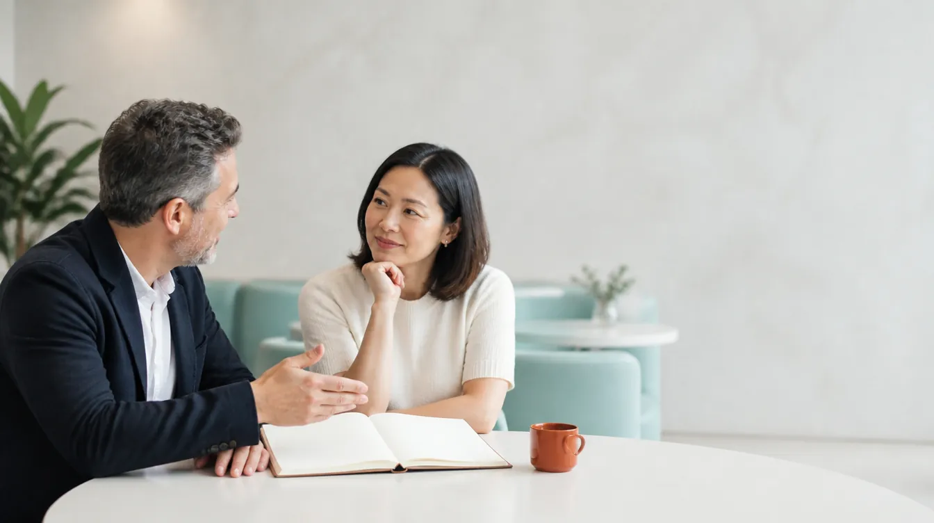 Two business partners in smart casual attire seated at a bright cafe table in relaxed negotiation conversation — one gesturing toward an open blank notebook