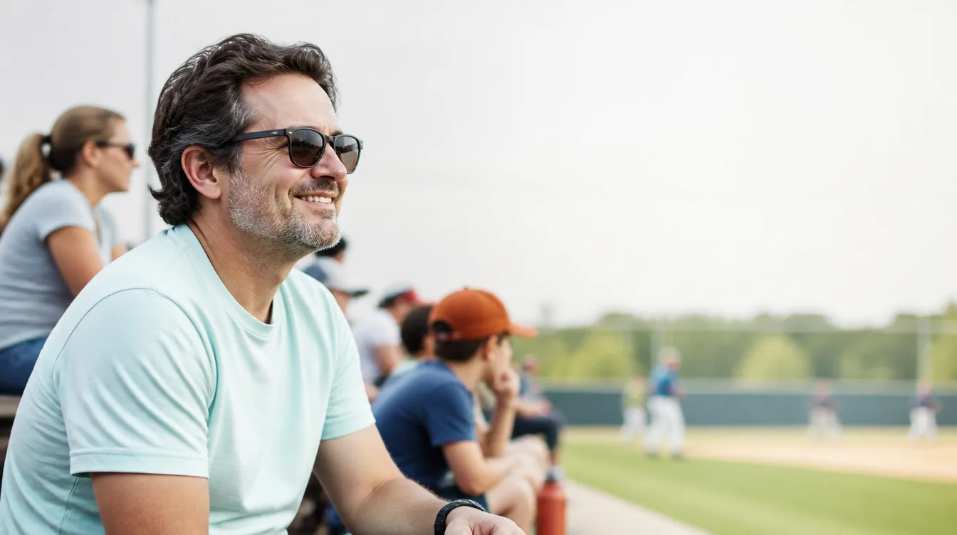 A parent in their 40s with natural skin sitting on outdoor bleachers watching a kids' baseball game, relaxed and smiling, casual weekend clothing with a pale mineral teal tone in